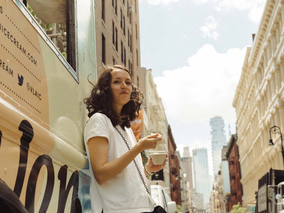 Woman in white t-shirt holding ice cream cup stands on New York street lined with tall buildings.