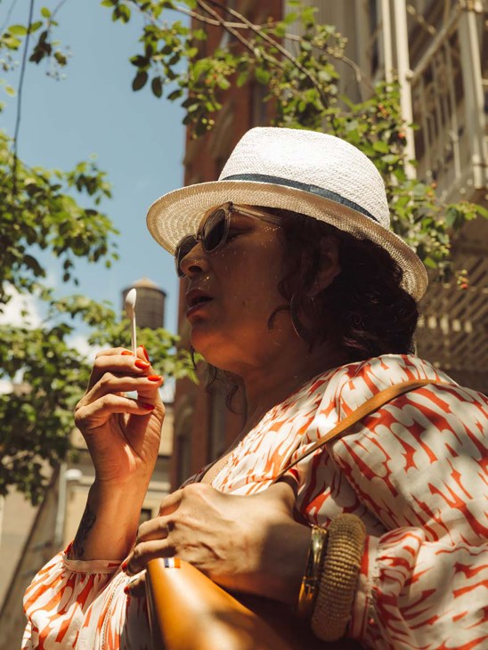 Woman in white and tan straw hat and red-patterned shirt holding an ice cream cone on a sunny urban street.