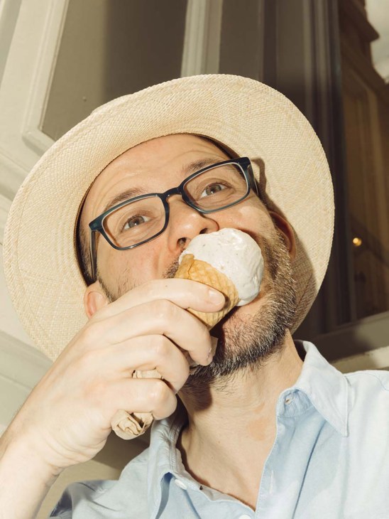 Man wearing glasses and a straw hat eating an ice cream cone with white topping.