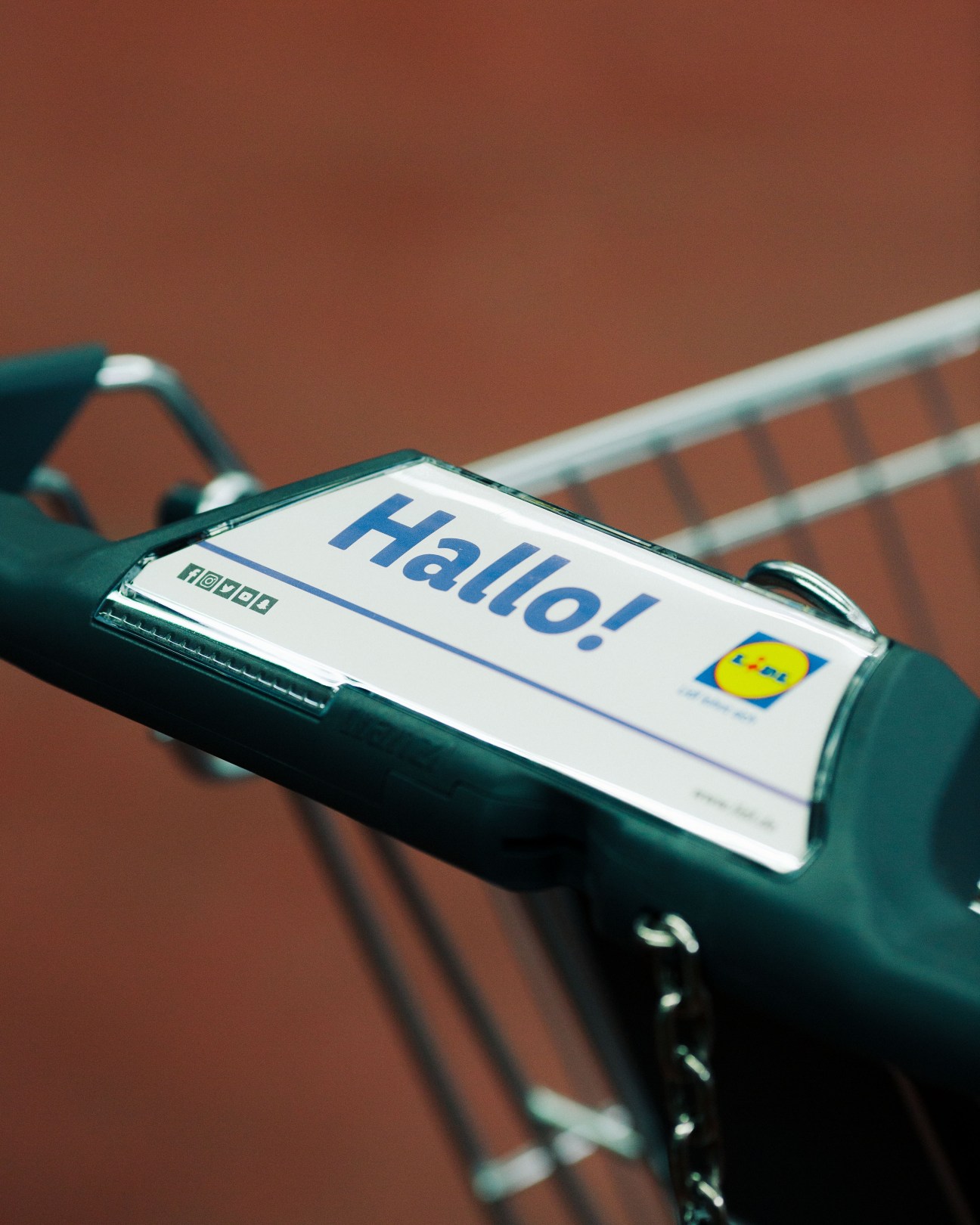 Close-up of a shopping trolley handlebar display showing a digital screen with 'Hallo!' greeting and Lidl logo against a reddish-brown background