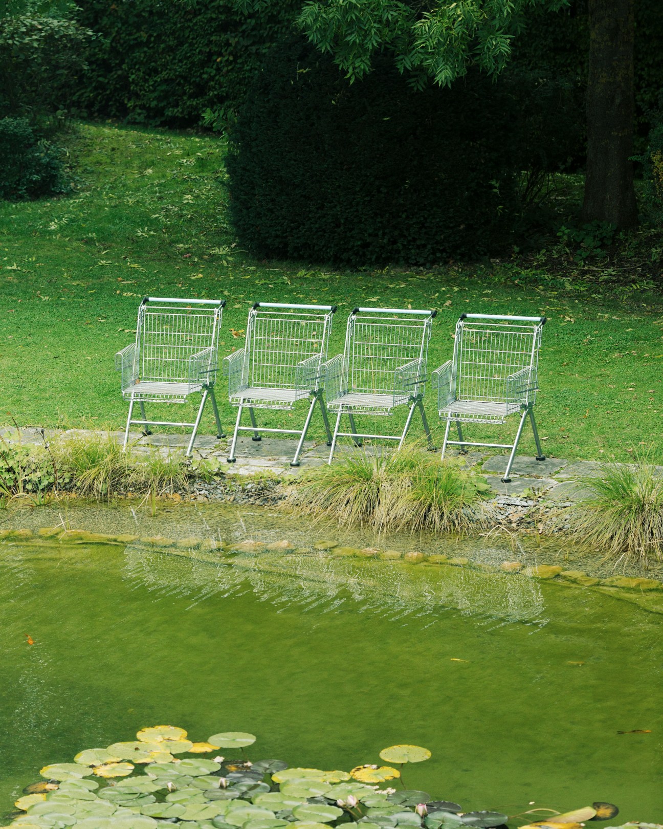 Four white metal shopping trolley-shaped chairs arranged by a pond at Wanzl headquarters