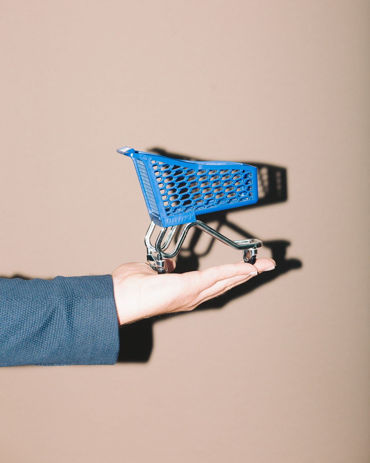 Hand holding a miniature blue shopping trolley with metal handle against a beige background.