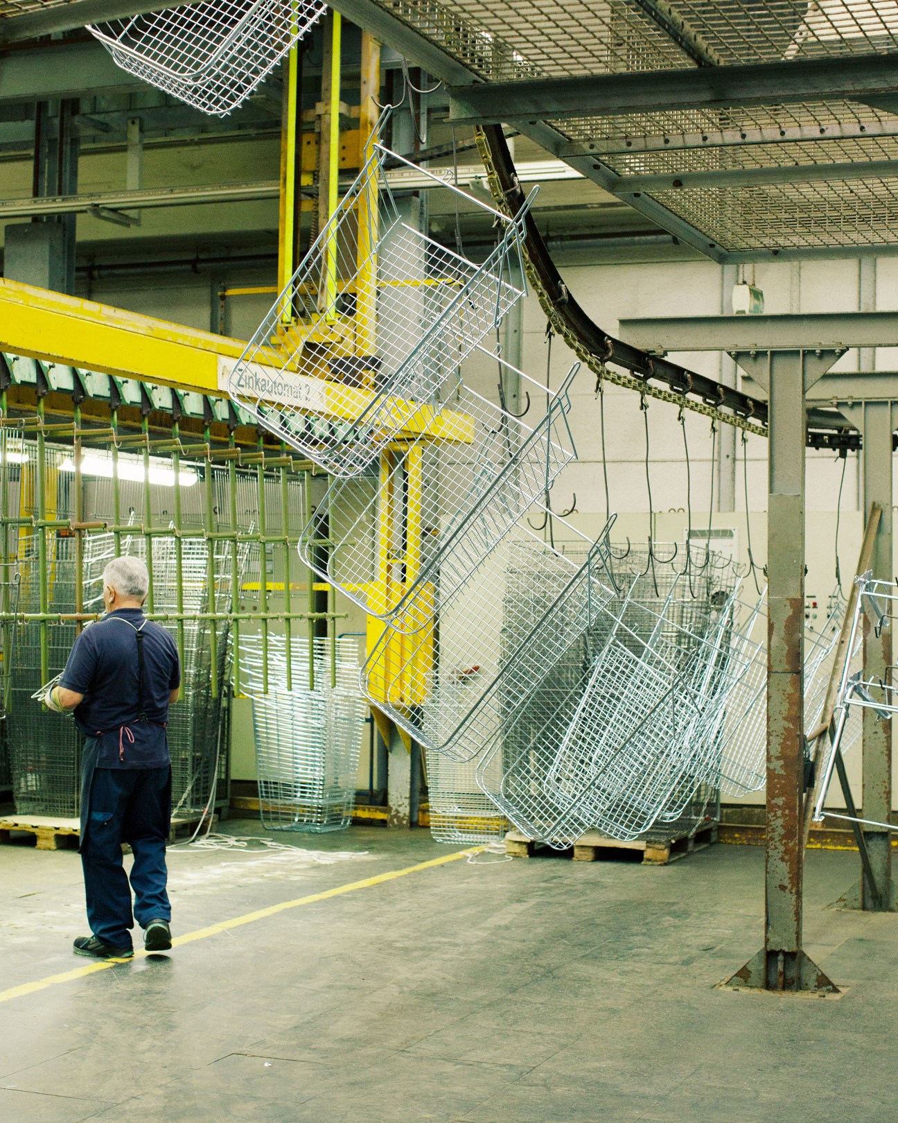 Shopping trolley wire baskets suspended on conveyor system above factory floor heading toward galvanizing process