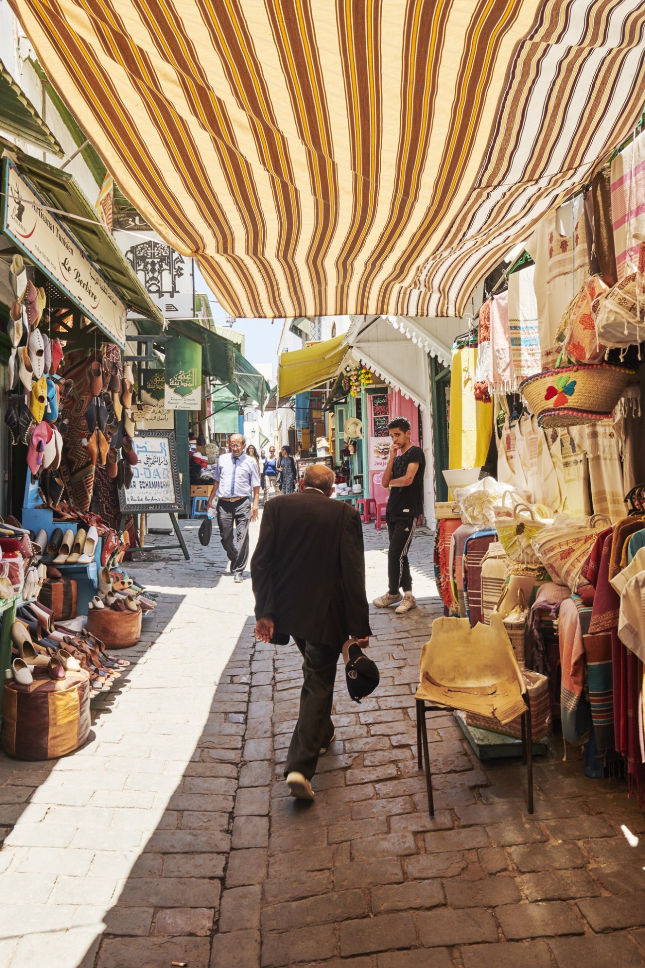 Man walking through a covered Tunisian souk market with striped awning and traditional craft shops.