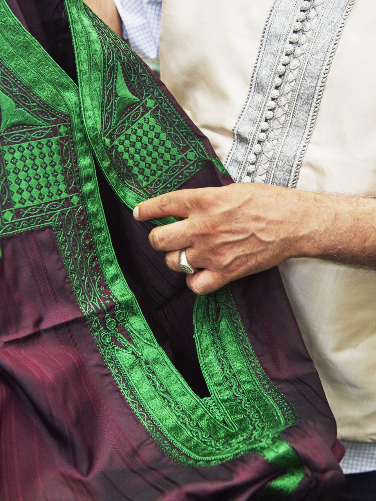 Close-up of intricate green embroidered trim on burgundy fabric held by a person's hand.
