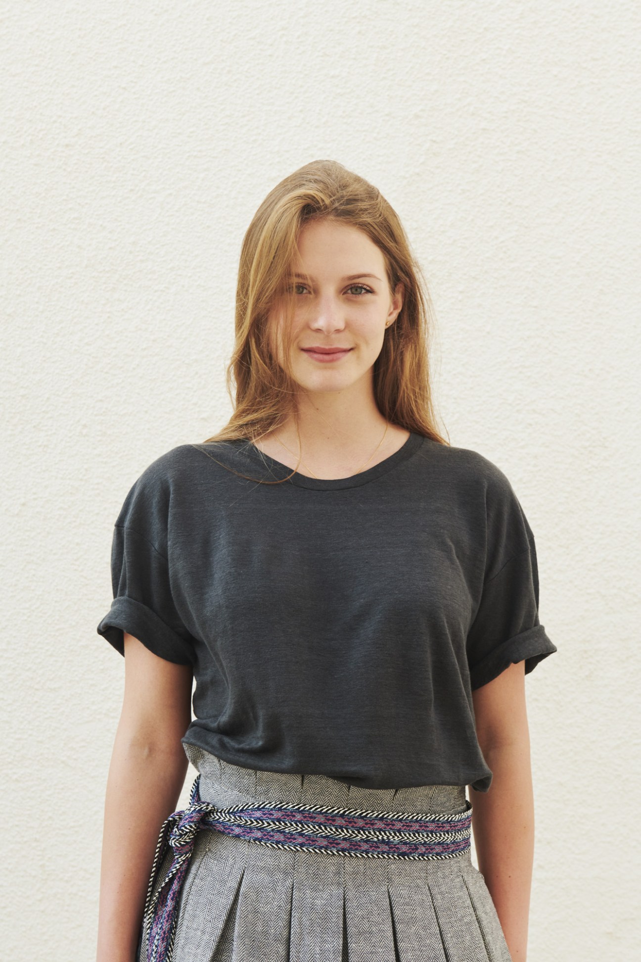 Woman wearing a dark t-shirt and gray pleated skirt with woven belt against white background.
