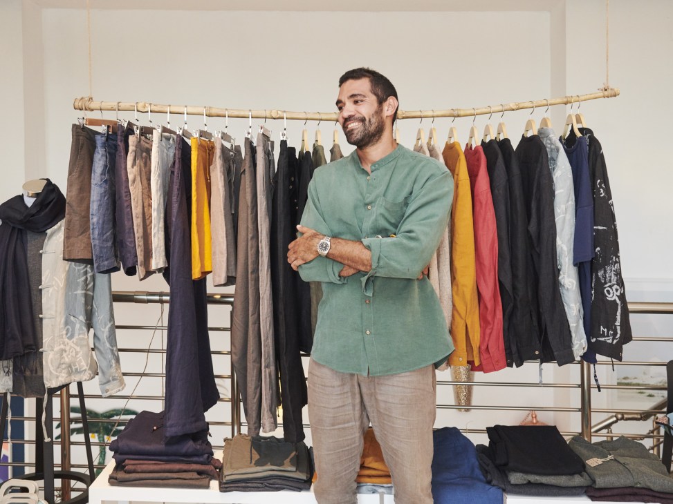 Aziz Kallel wearing a light green shirt and beige trousers from his Samaka clothing line, standing in front of a clothing rack.