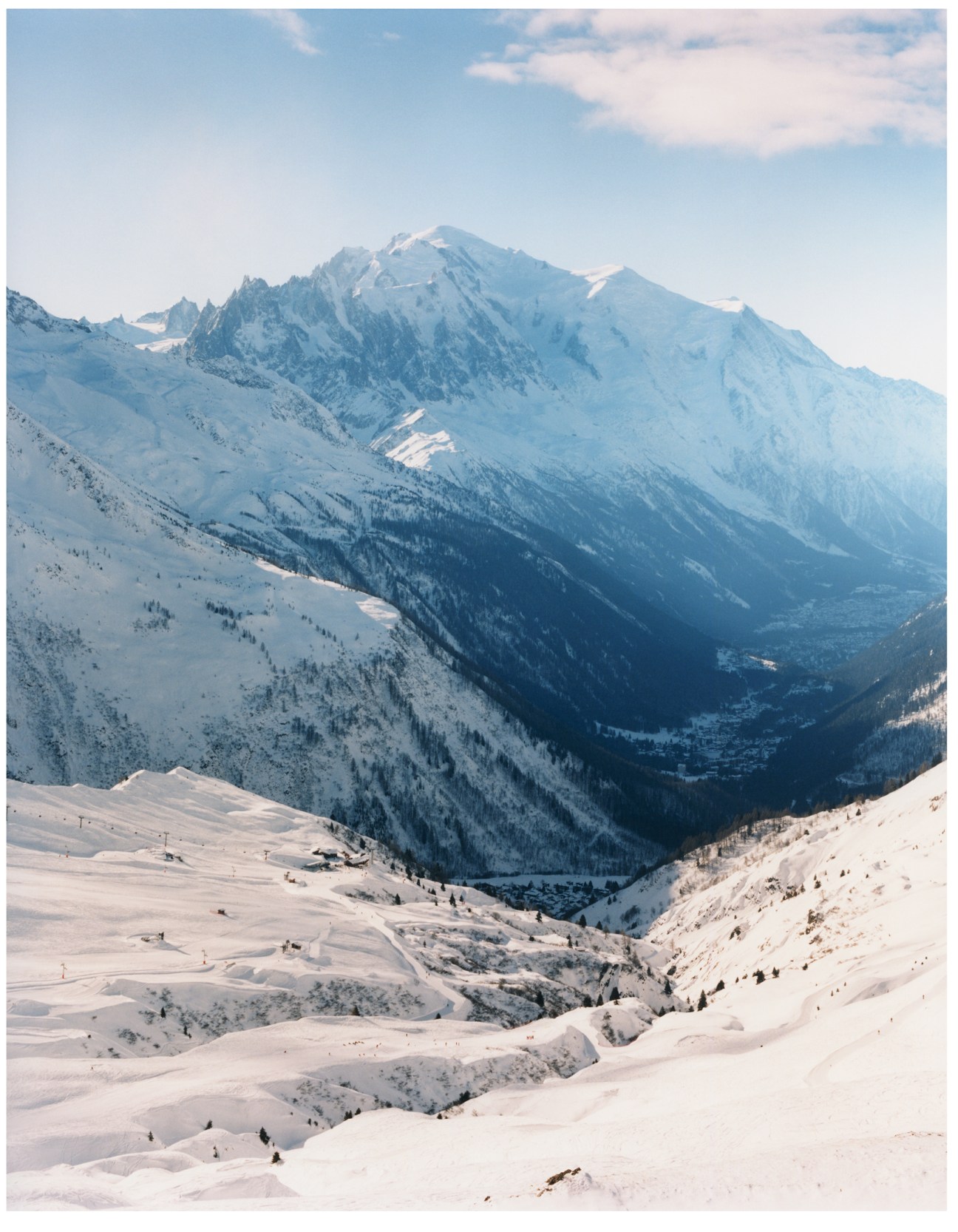 Snow-covered Mont Blanc mountain rises above a forested valley with ski slopes visible in the foreground.