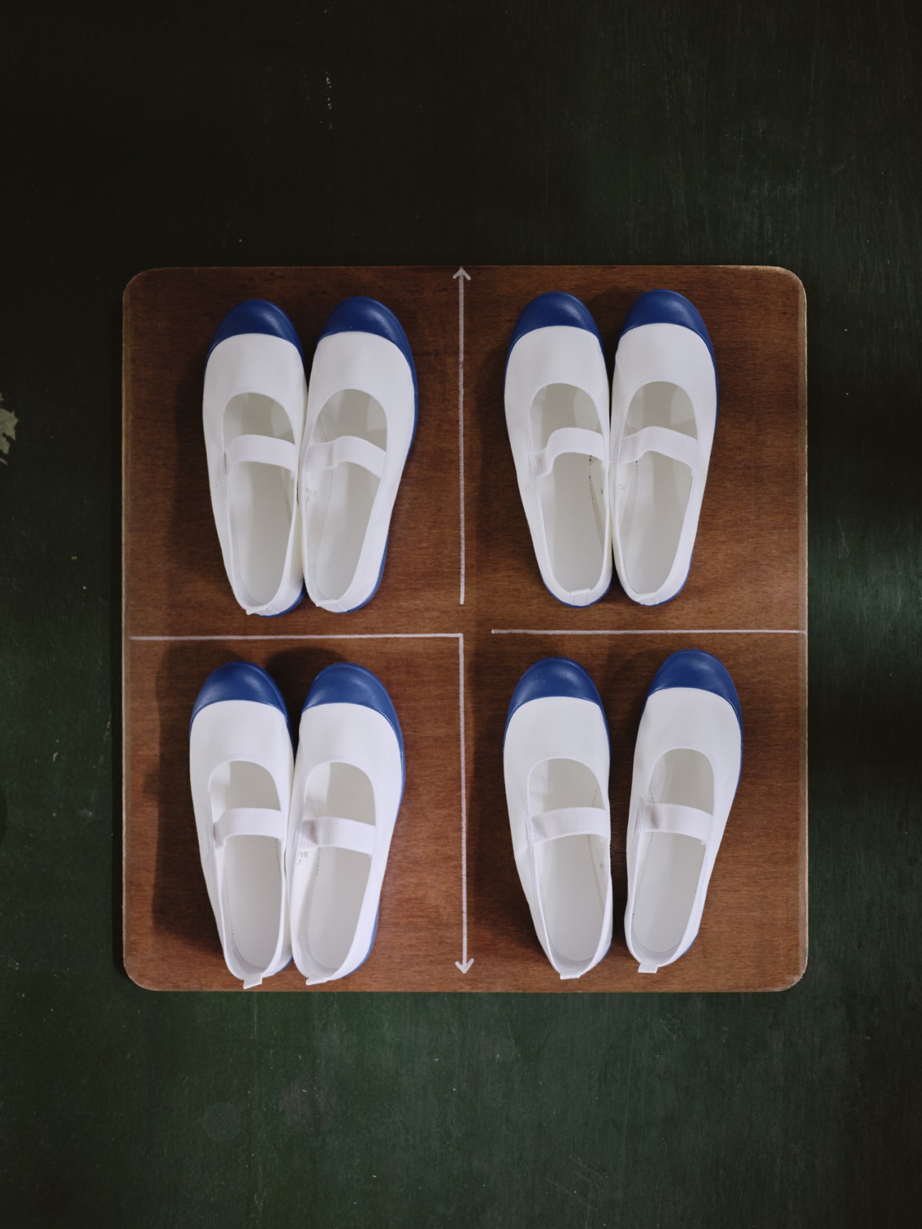 Four pairs of white canvas slip-on plimsolls with blue heels arranged on a wooden board.