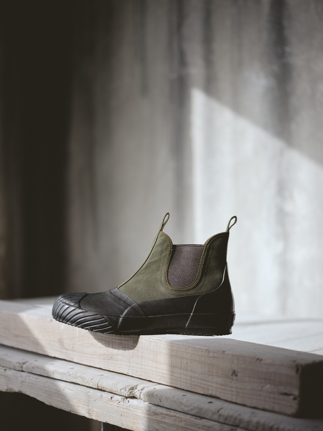 Moonstar's canvas and rubber Chelsea boot with olive-green canvas upper and black rubber sole displayed on weathered wood.