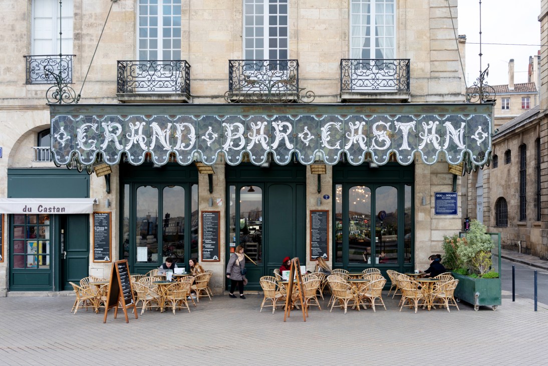 Le Grand Bar Castan storefront with ornate signage, green doors and wicker chairs on the terrace in Bordeaux.