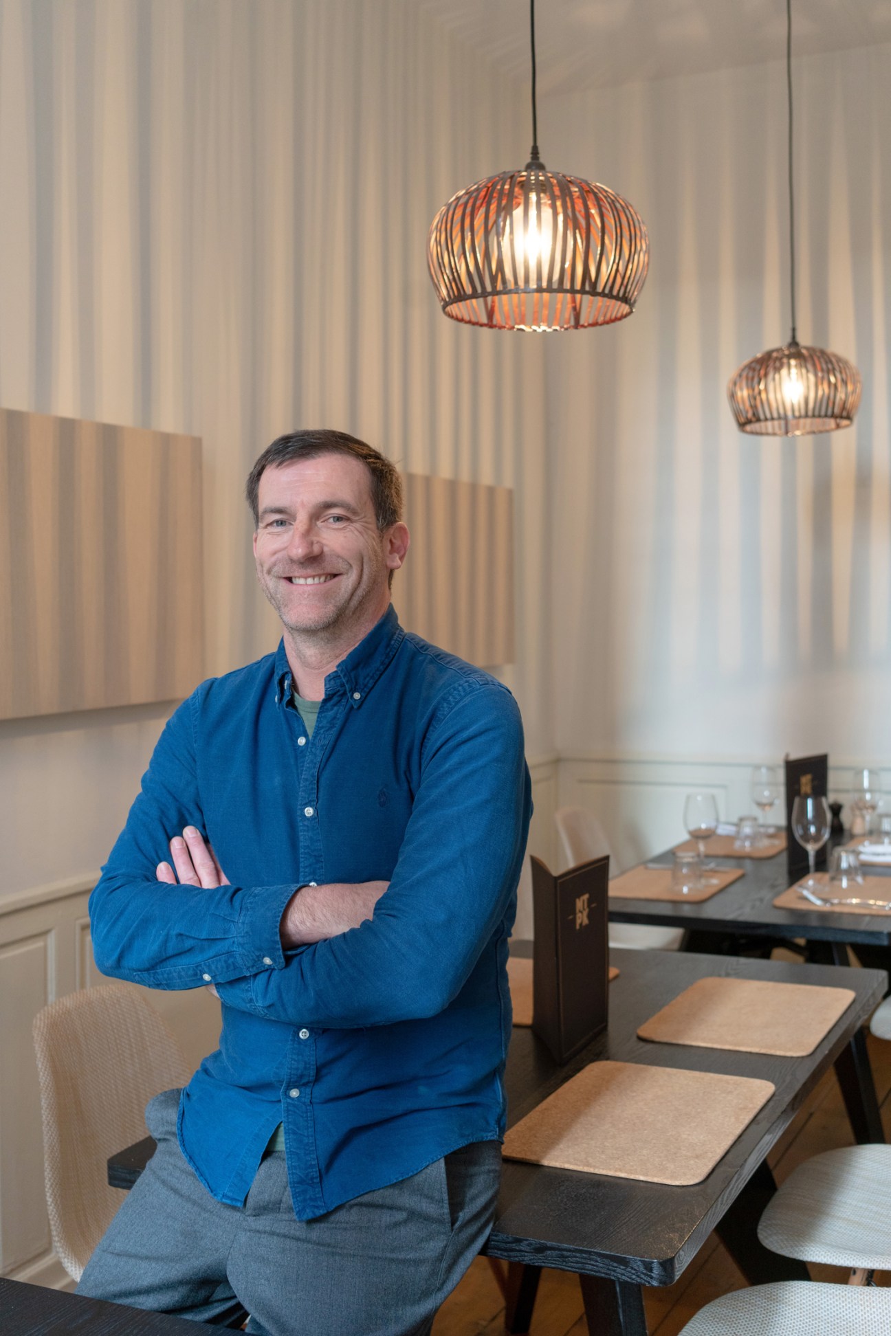 Tim Remi sits in his restaurant wearing a blue denim shirt, with pendant lights above and dining tables visible in the background.