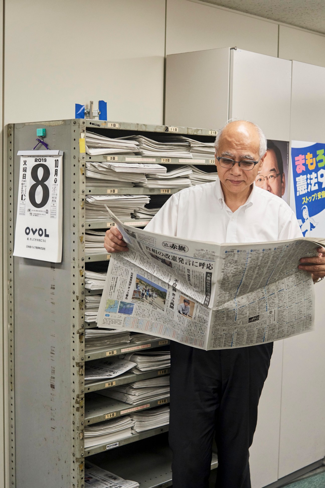 Yoji Kogiso, editor in chief of JCP newspaper Shimbun Akahata, reading a Japanese newspaper in an office with shelves of archived papers behind him.