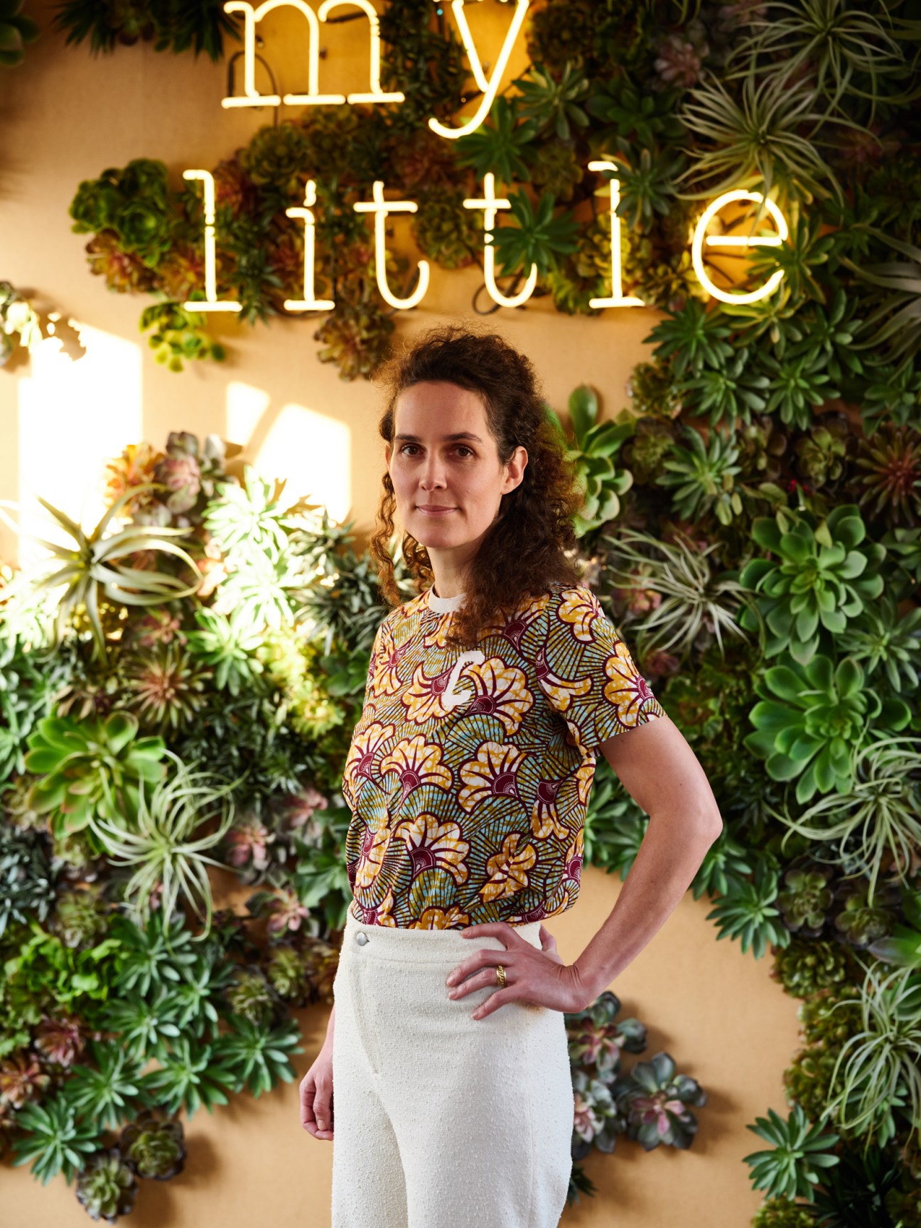 Céline Orjubin, co-founder of My Little Paris, poses in front of a living wall of green plants under a neon 'my little' sign