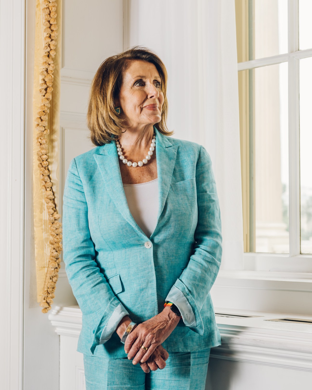 Woman in teal blazer and pearl necklace standing by window in formal interior setting
