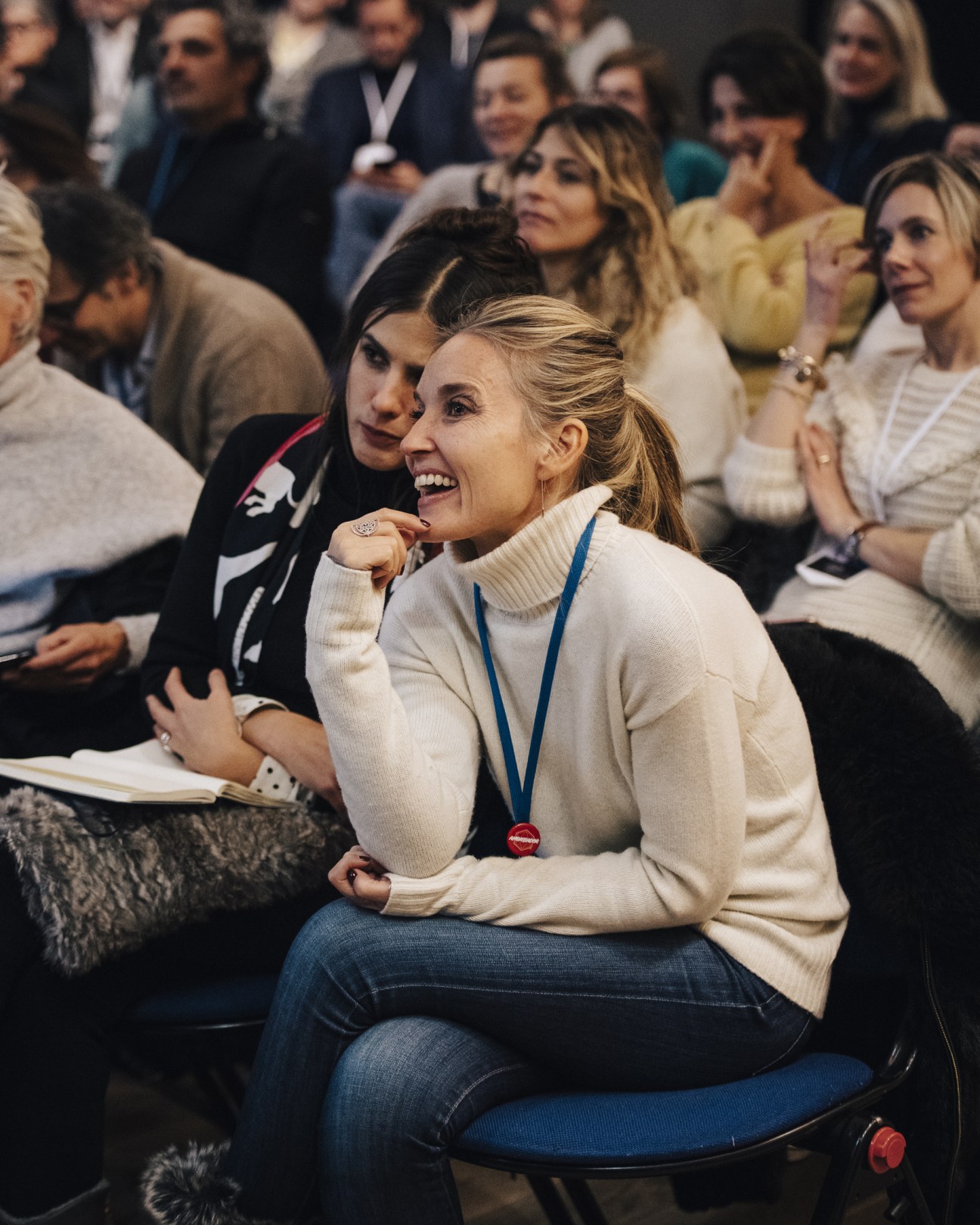 A smiling woman in a cream sweater with a lanyard sits among other attendees at a Les Napoleons workshop.