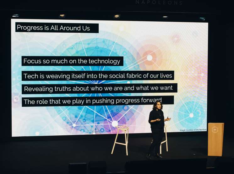 A woman speaker gestures while presenting on stage at a Les Napoleons event with a large screen displaying progress-related quotes behind her.
