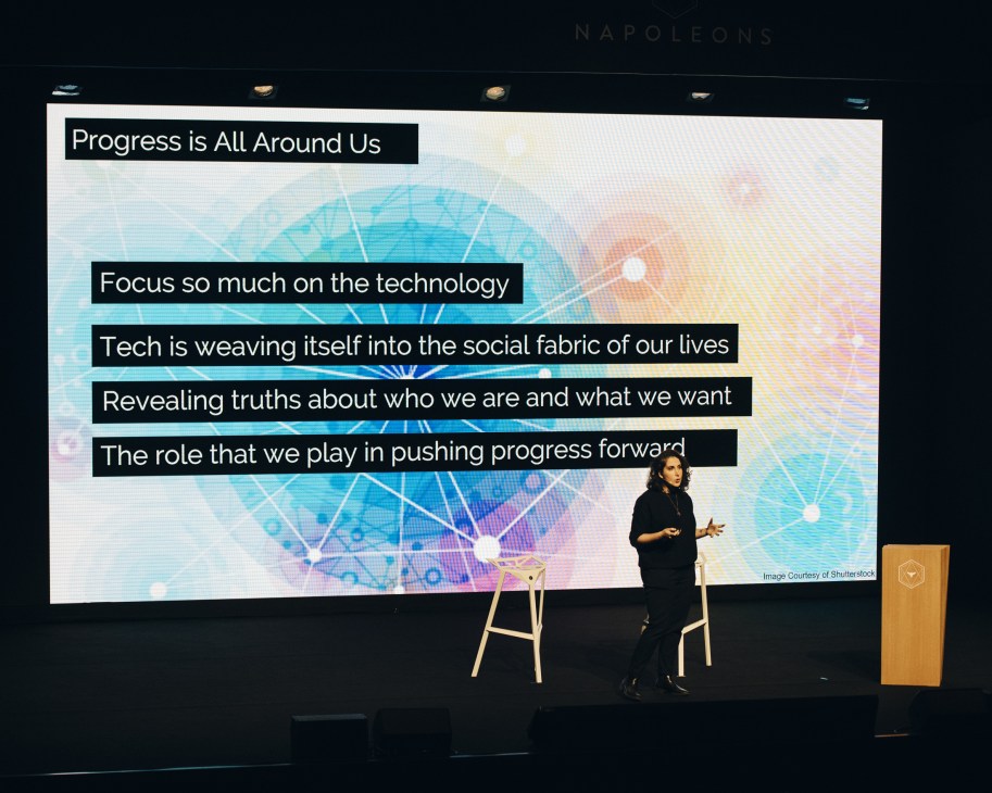 A woman speaker gestures while presenting on stage at a Les Napoleons event with a large screen displaying progress-related quotes behind her.