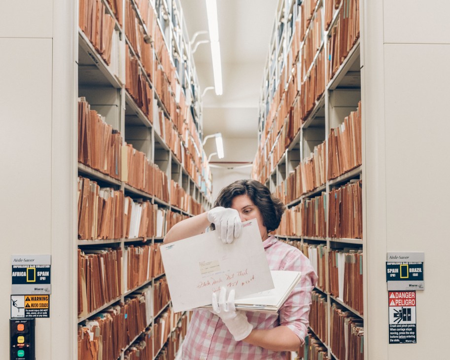 Archivist Rebecca Dupont wearing white gloves examines a document in National Geographic's basement archives lined with brown filing boxes.