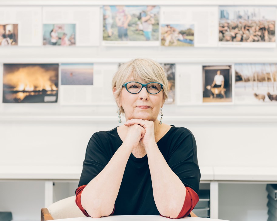 Director of photography Sarah Leen poses with hands clasped in front of a wall displaying various photographs.