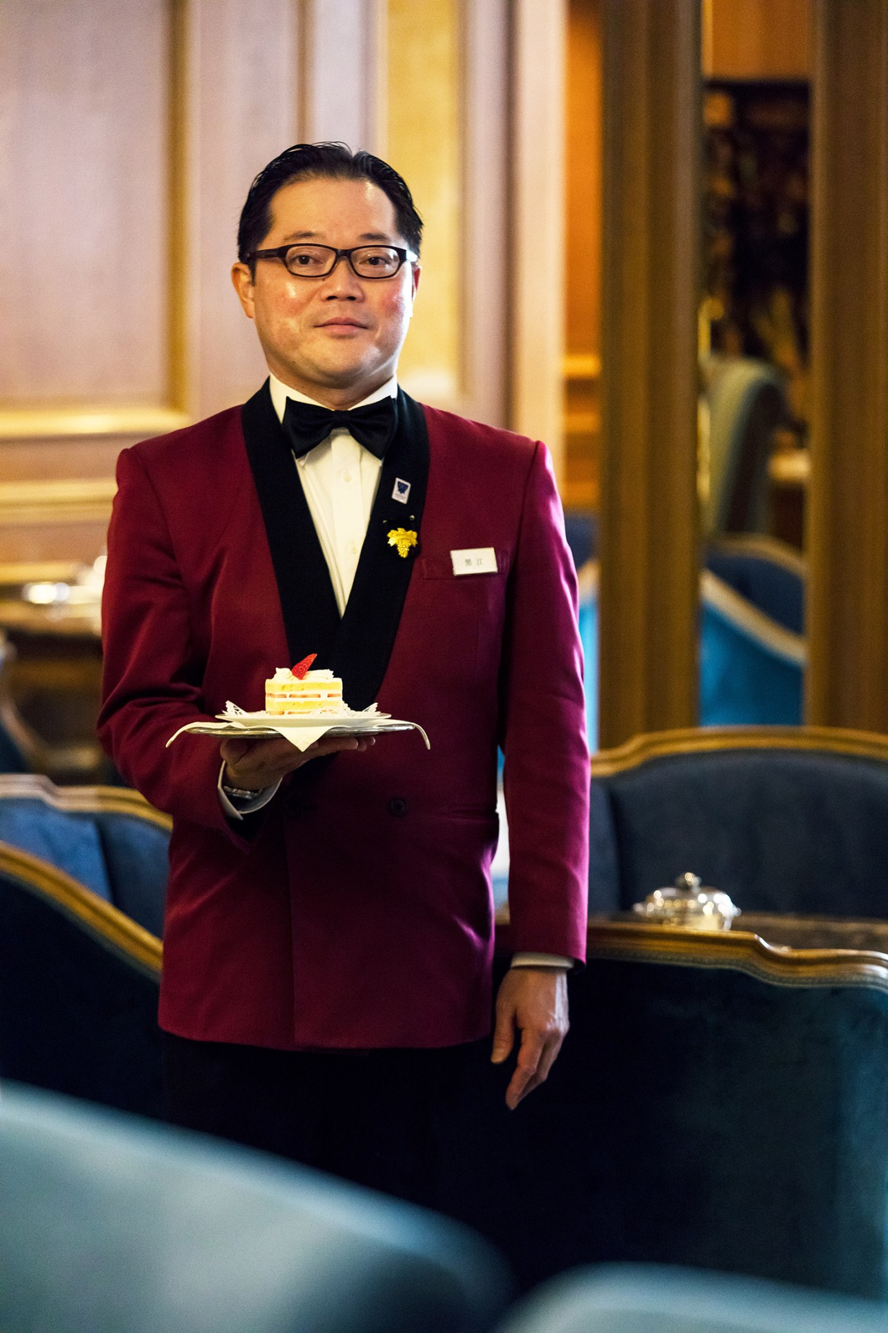 Hotel staff member in maroon jacket and bow tie serving a dessert plate in the Hotel New Grand's lobby lounge.