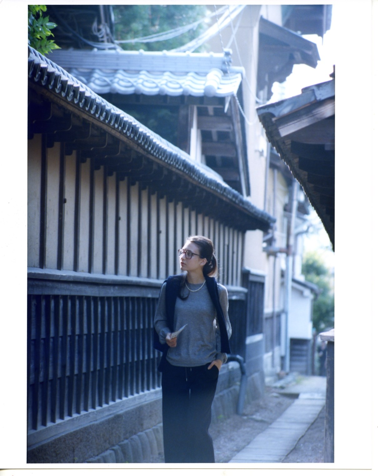 Model wearing Drawer jumper and Bottega Veneta glasses stands in traditional Japanese street with wooden architecture and curved roof tiles.
