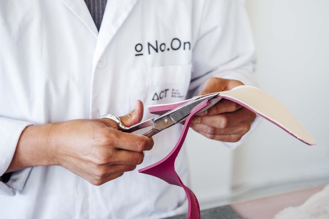 Person in white No.On lab coat holding scissors and pink fabric while cutting material for shoe production.