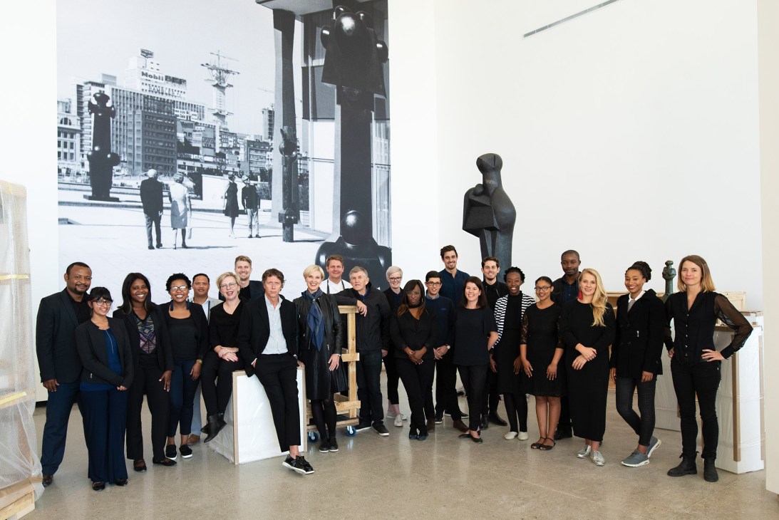 A diverse team of approximately 25 people standing together in a gallery space with artworks and sculptures visible in the background.