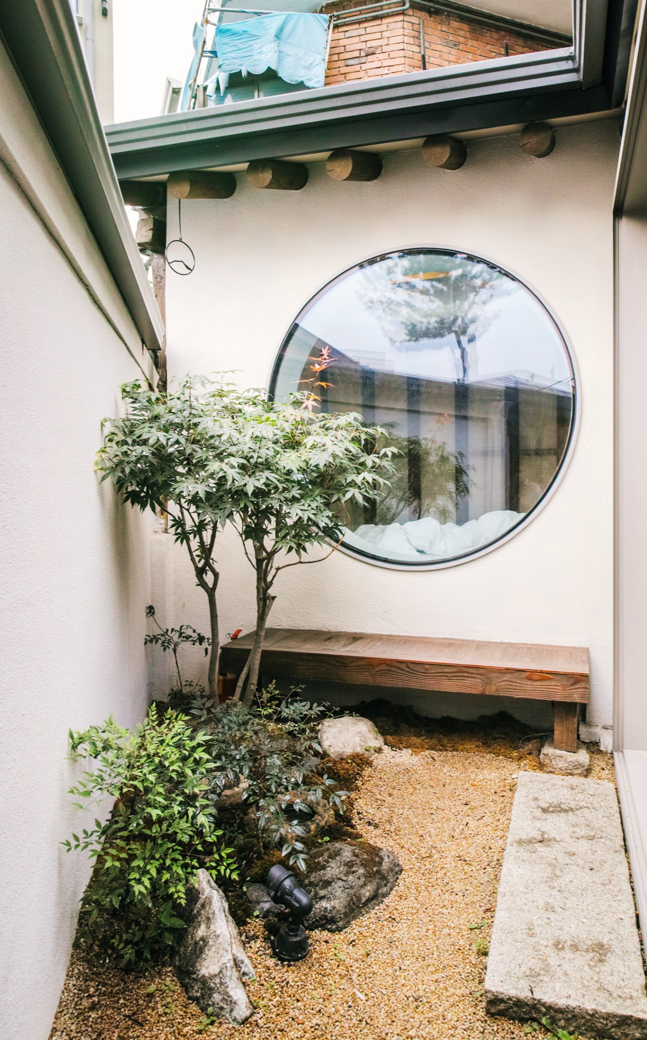 Courtyard at Nuwa with circular window, small tree, bench, and gravel ground in Seoul hanok.