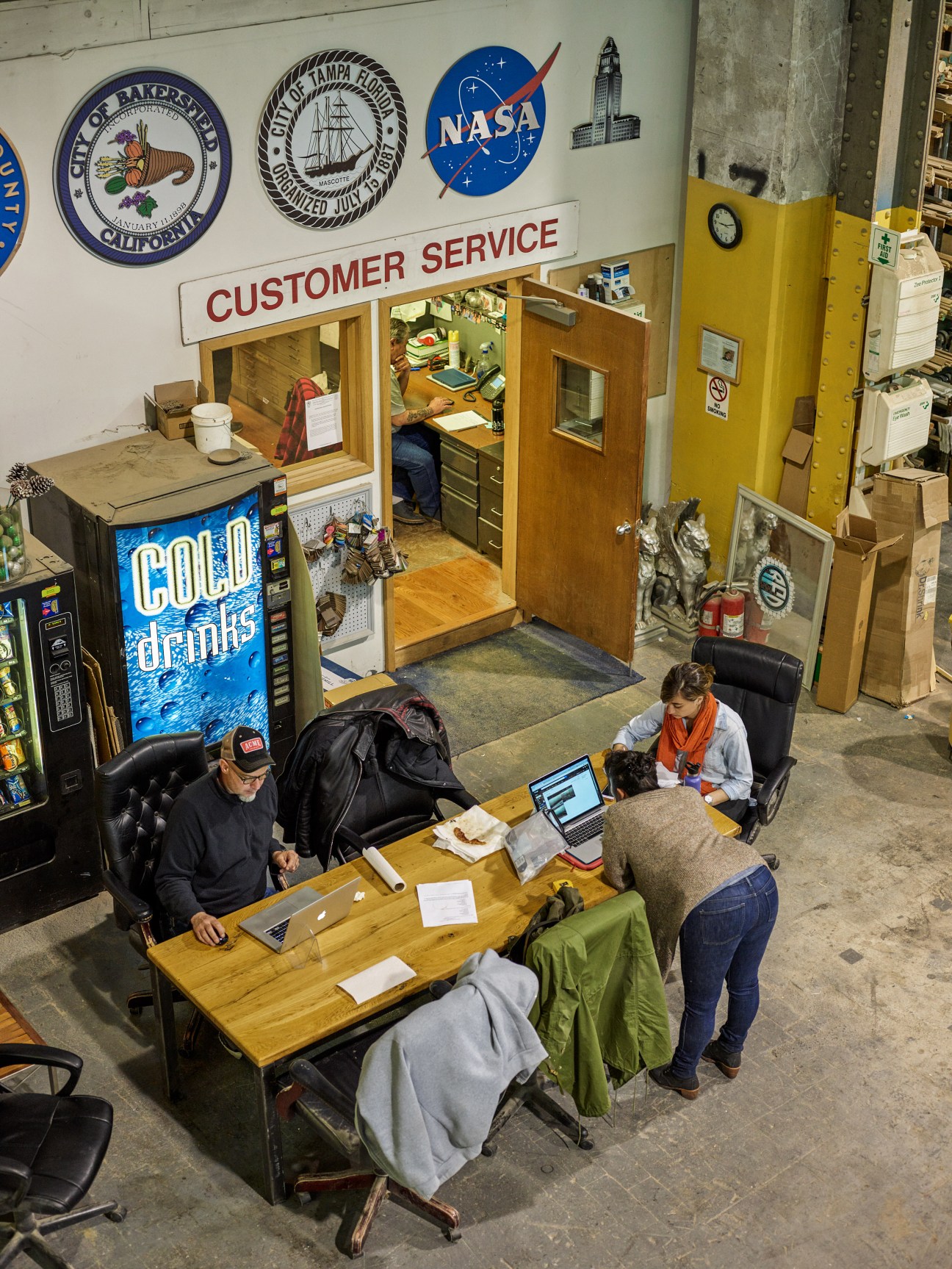 Office workspace at Stiegelbauer Associates with employees at desk and prop set pieces including customer service booth with NASA and city seals.