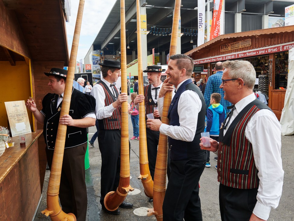 Swiss musicians in traditional clothing playing large wooden alphorns at the Olma fair.