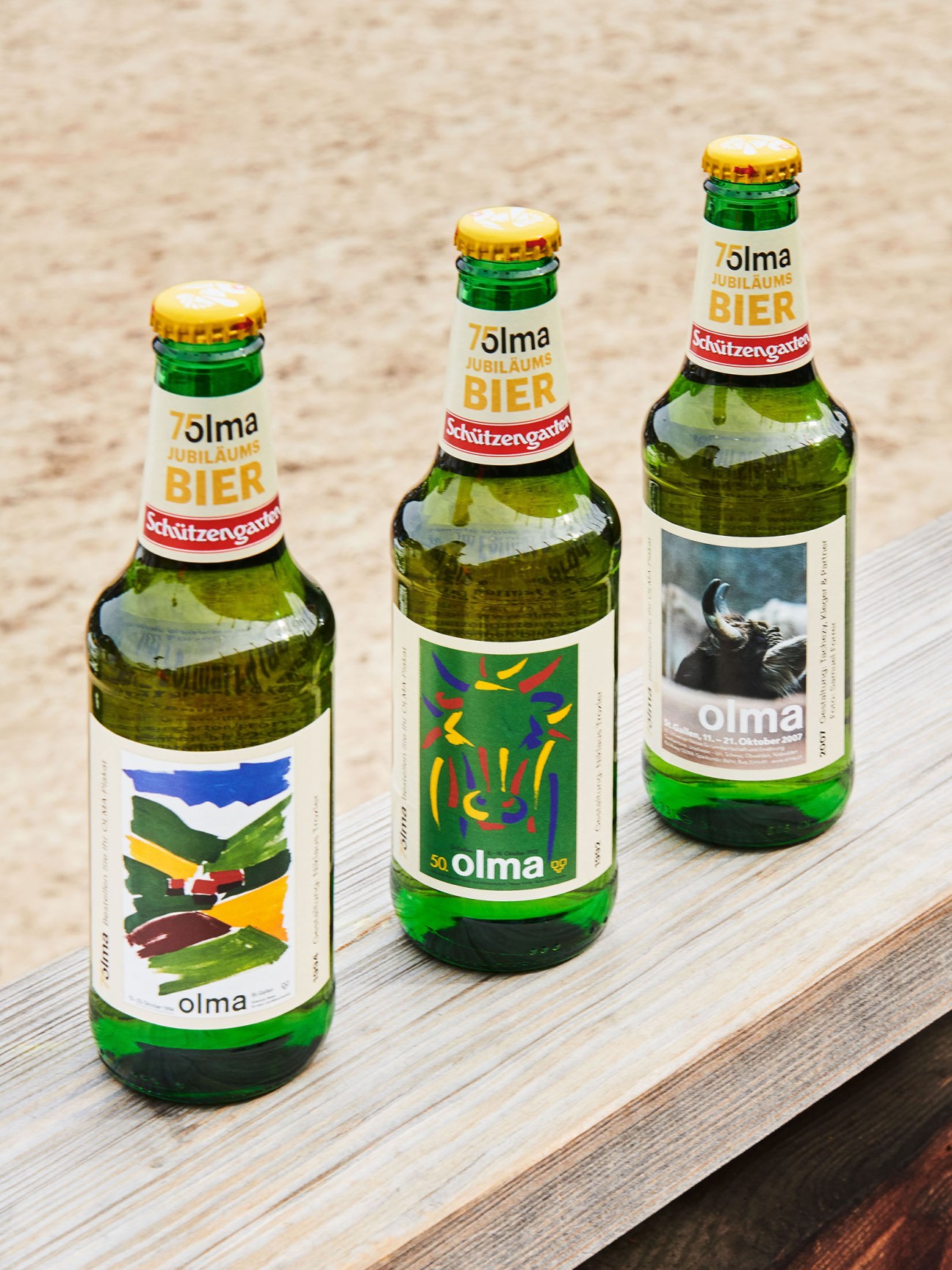 Three green glass bottles of Schützengarten Olma Jubiläums Bier with gold caps on a wooden surface.
