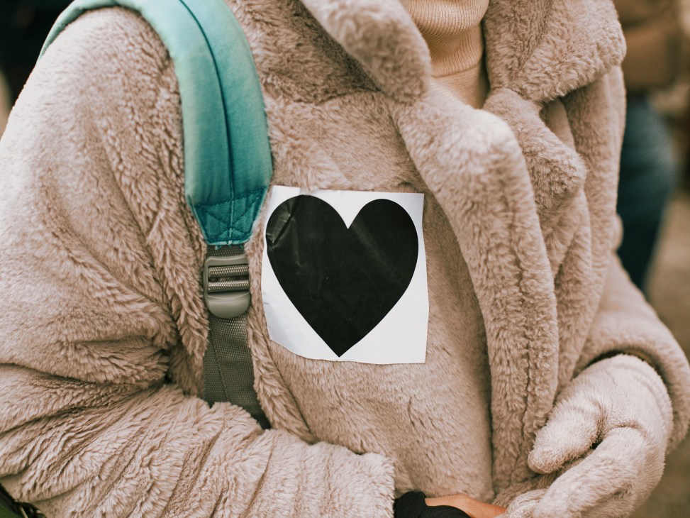 Person wearing beige fleece jacket displaying black heart symbol on white paper pinned to chest.