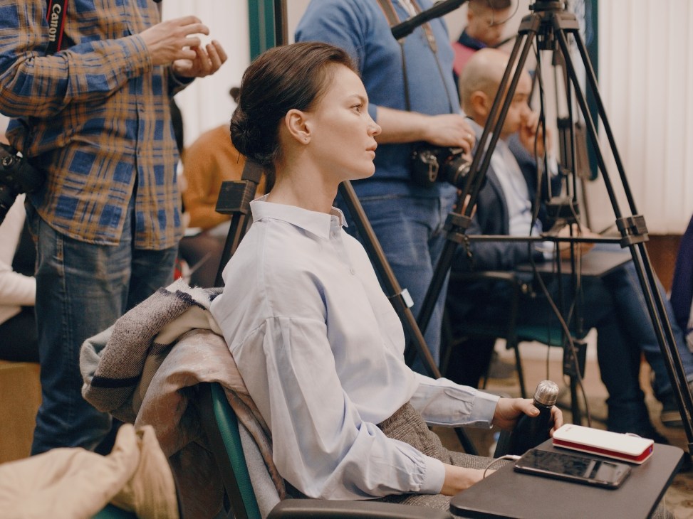 Woman in white shirt speaking at Open Russia press conference surrounded by journalists and camera equipment.
