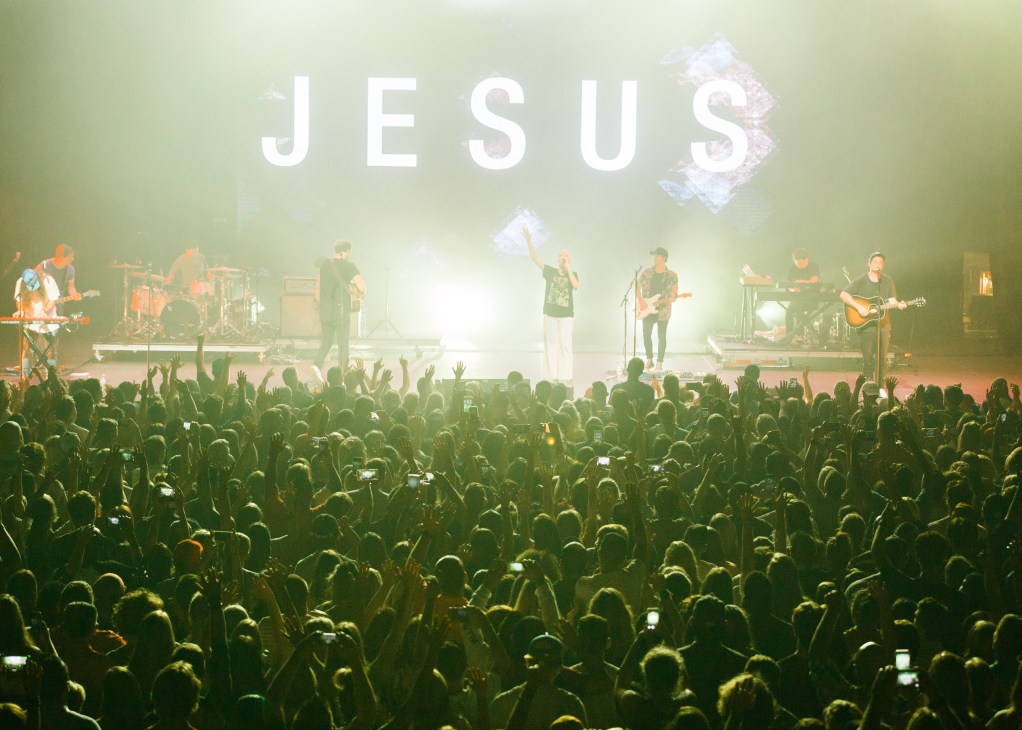 Hillsong United performing on stage with large crowd and 'JESUS' text projection above.