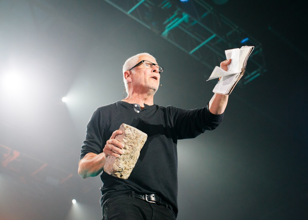 Preacher Louie Giglio speaking at the Outcry Festival, holding papers and a brick while addressing the audience.