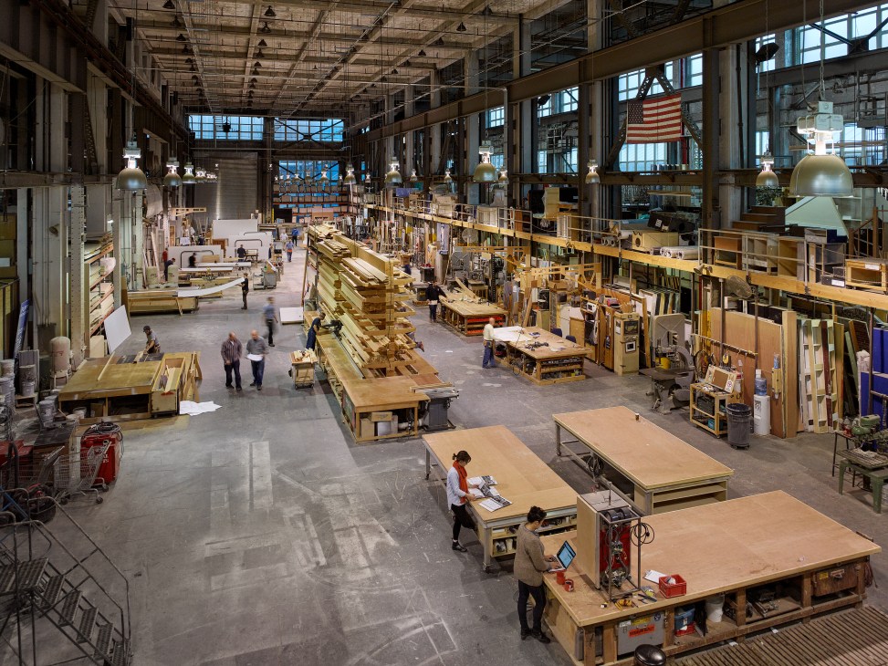 Aerial view of Stiegelbauer Associates' industrial workshop in Brooklyn Navy Yard with workers, wooden set pieces, and construction materials.