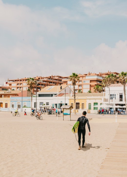 Surfer in wetsuit carrying yellow-green board walking on Patacona Beach toward colorful low-rise buildings and palm trees.