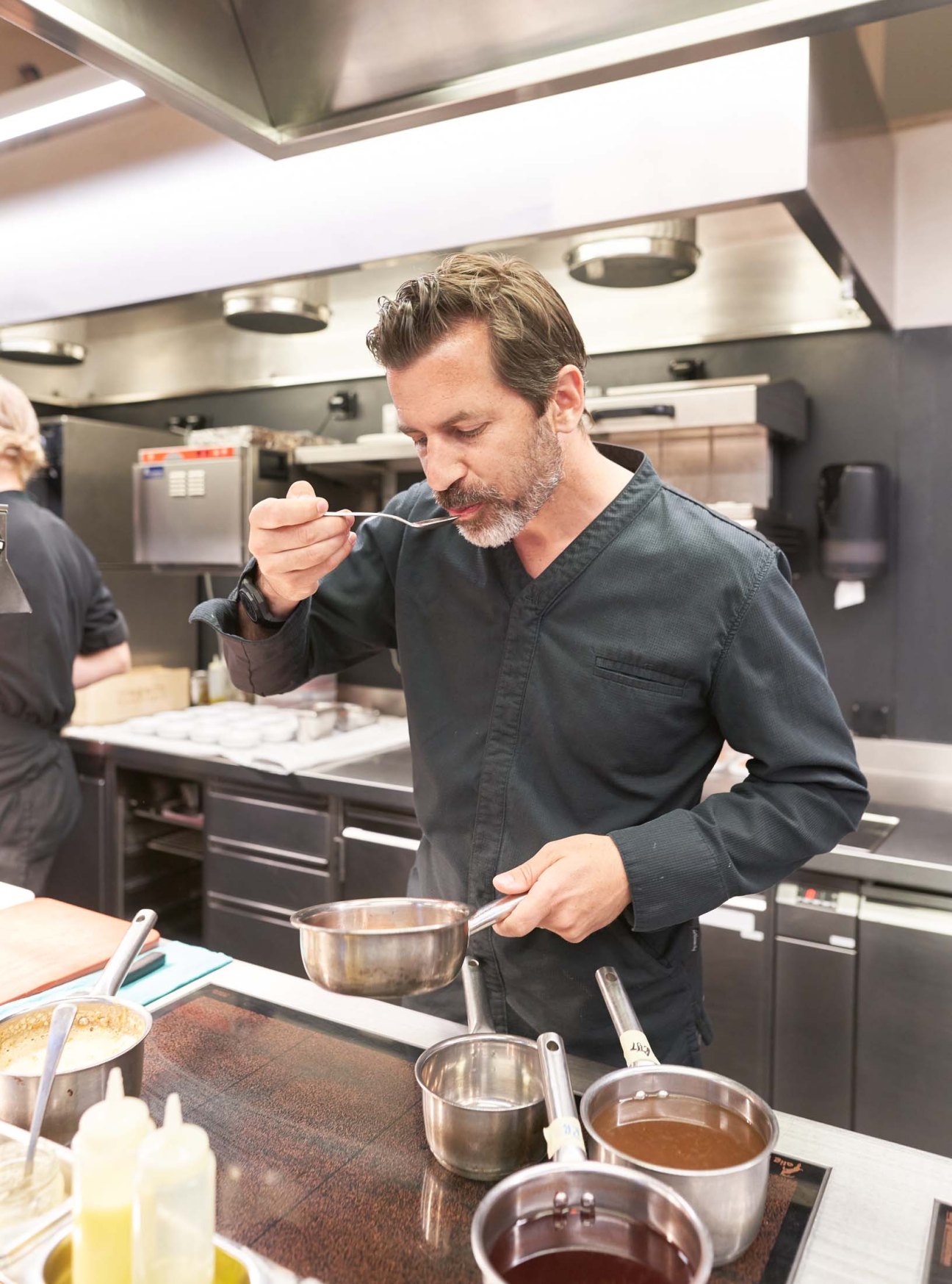 Chef Andreas Caminada in dark chef's whites tasting food in a professional kitchen