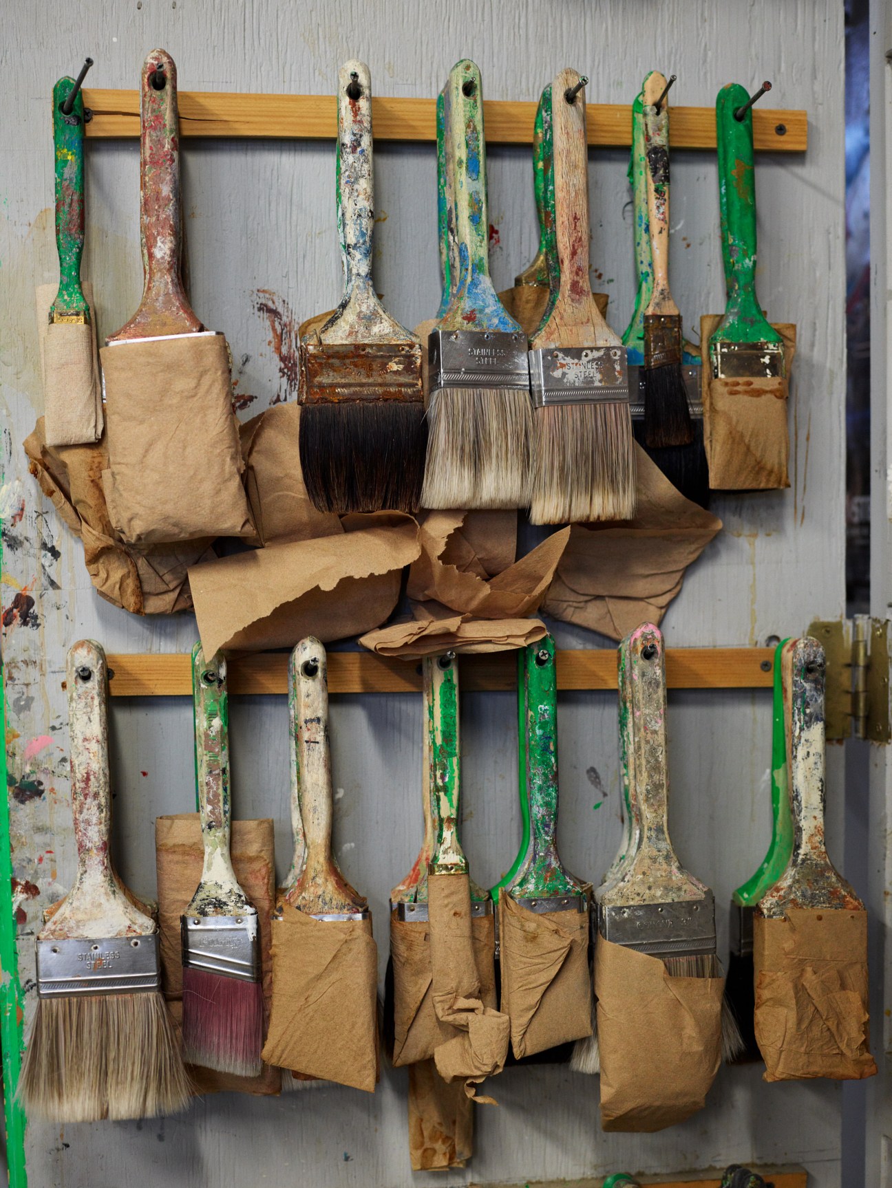 Paint brushes with colorful handles and worn paper sheaths hanging on wooden racks.
