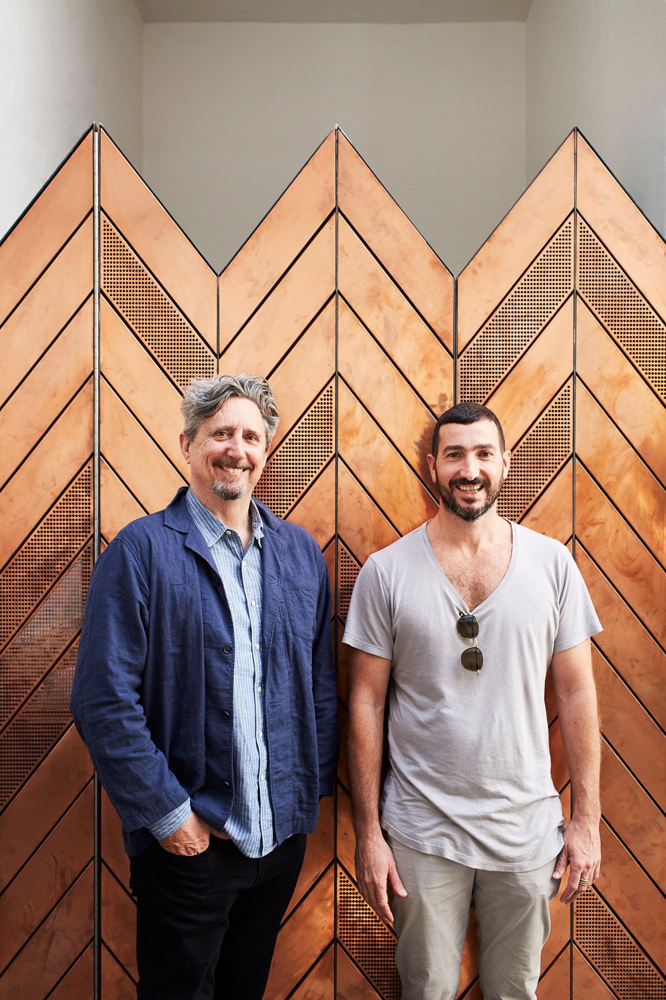 Co-founders Mark Dundon and Russell Beard stand in front of a copper geometric wall feature at Paramount House Hotel.