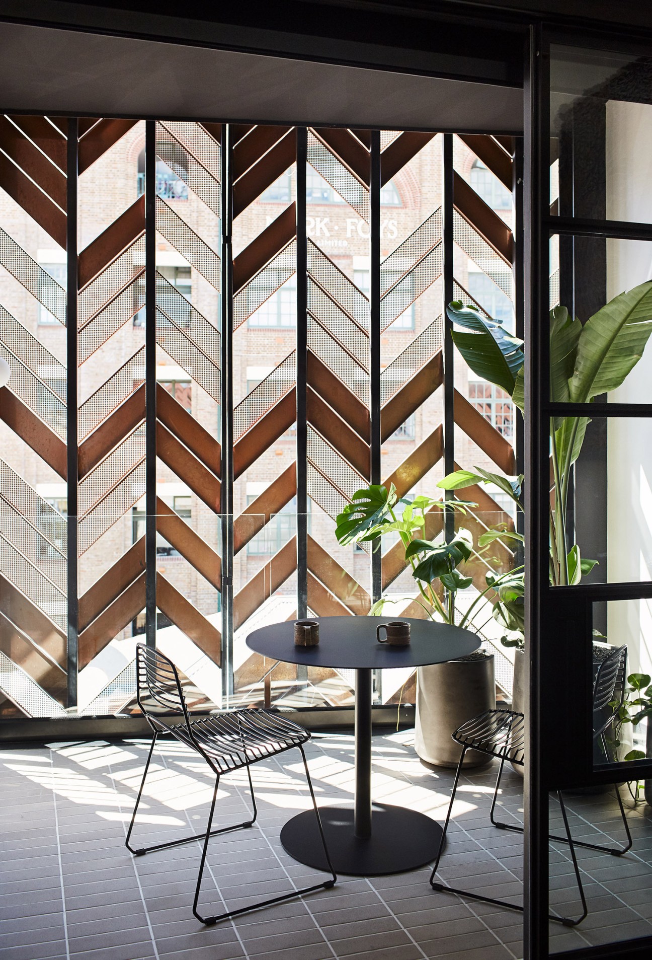 A private patio with a black round table, wire chairs, and green plants behind geometric wooden screens.