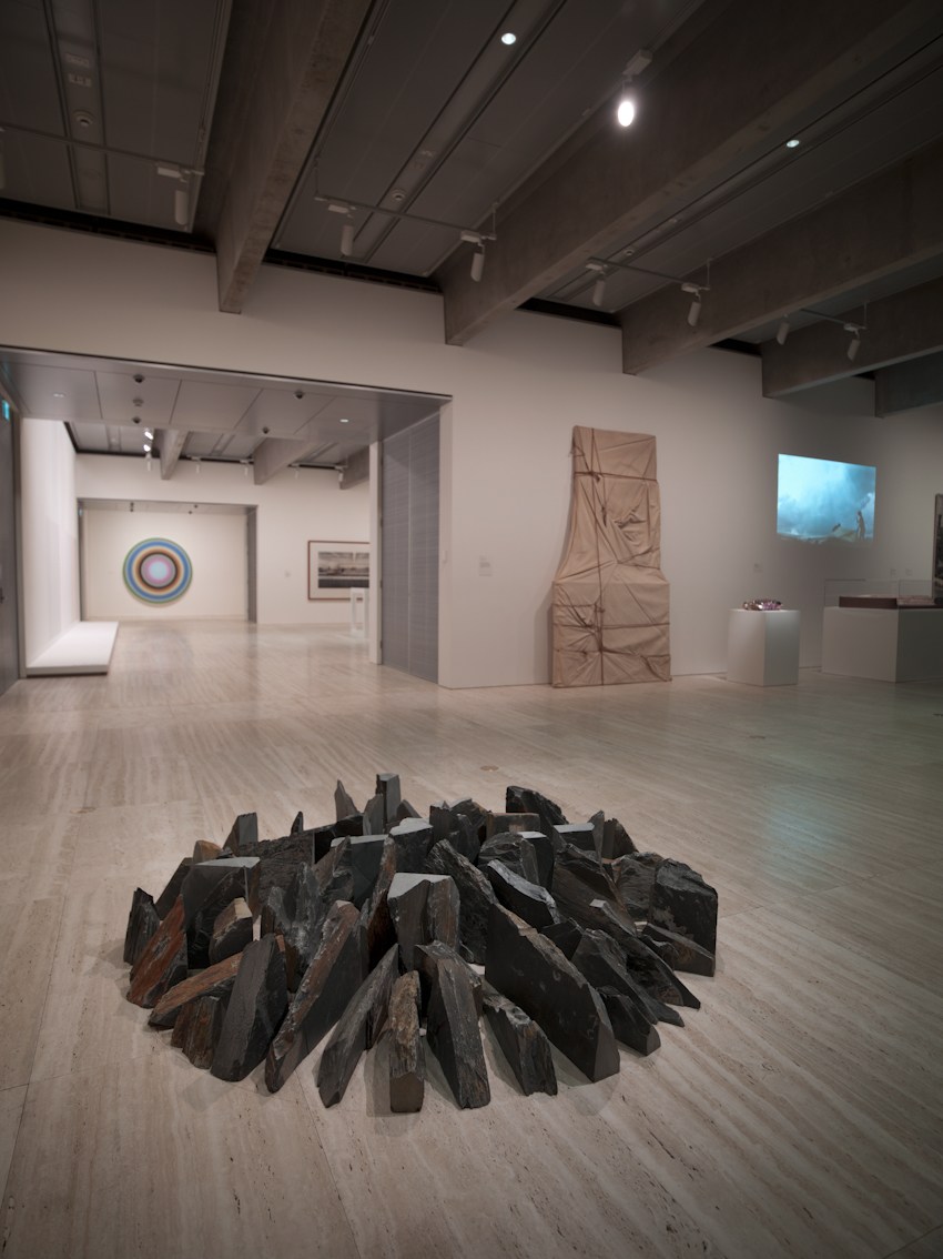 Gallery interior displaying Richard Long's 'Spring Showers Circle', a circular arrangement of dark wooden pieces on a wooden floor, with other contemporary artworks visible in the background.