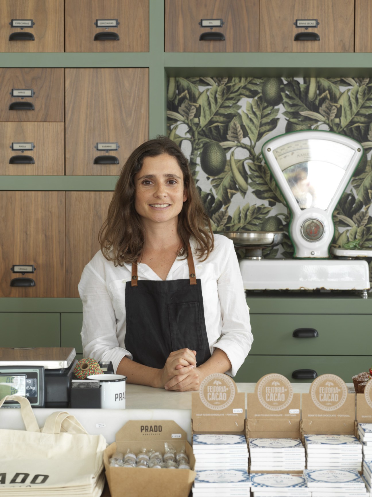 Grocer Marta Soares stands behind the counter at Prado Mercearia with wooden storage drawers and a vintage scale behind her.