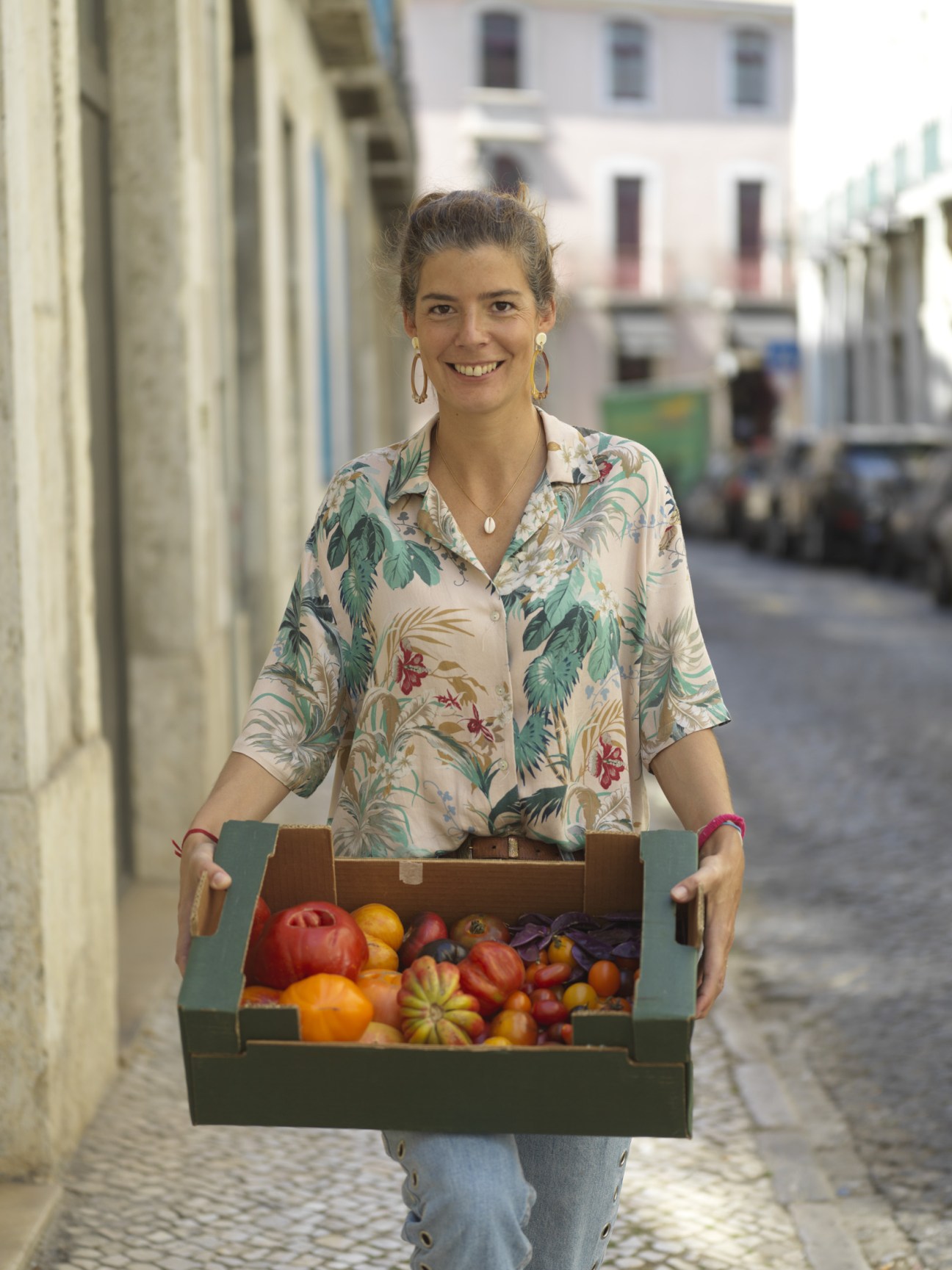 Woman in floral shirt holding a wooden crate of colorful tomatoes in a Lisbon alleyway.