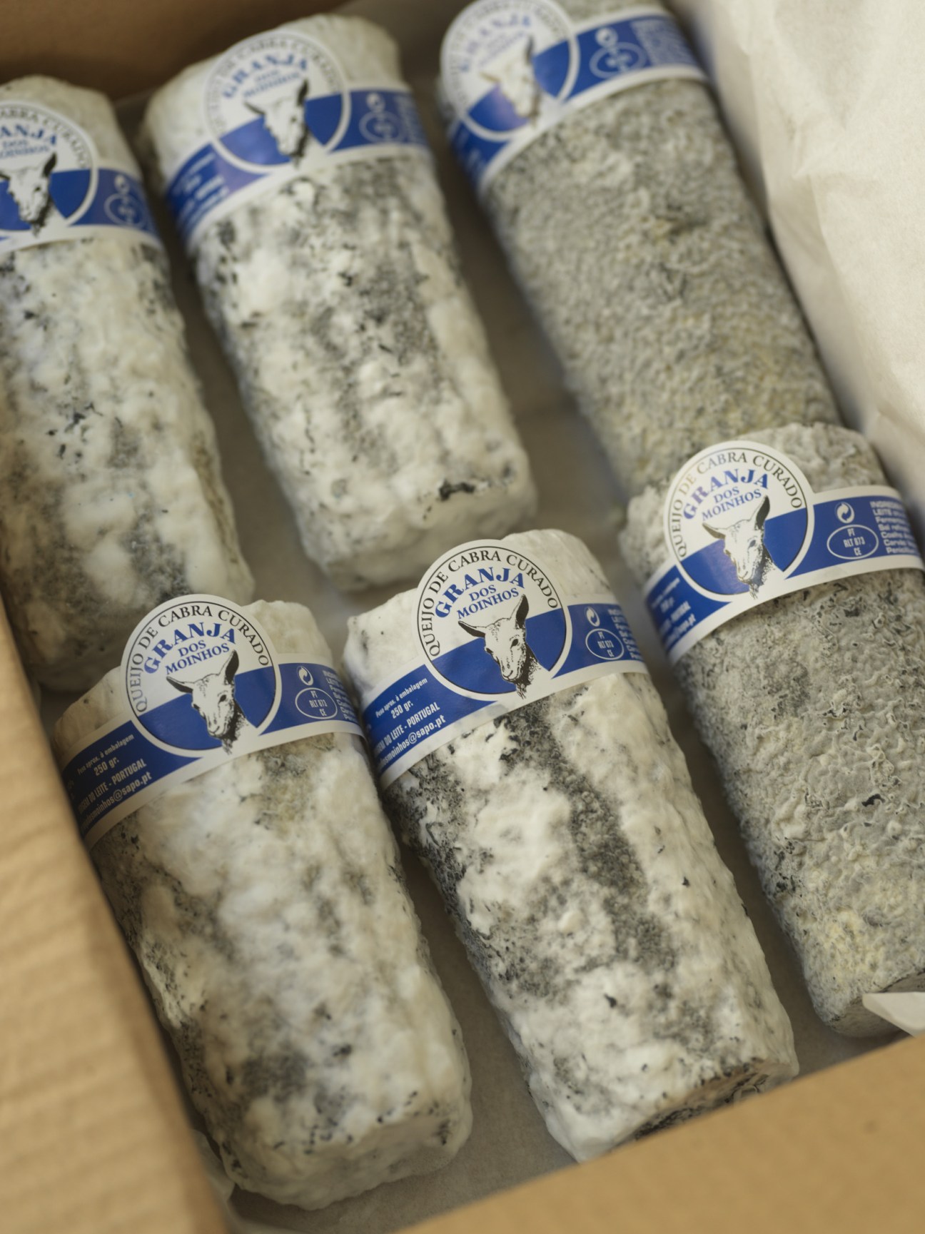 Rolls of goat's cheese with blue and white labels from farmer Adolfo Henriques displayed in a box.