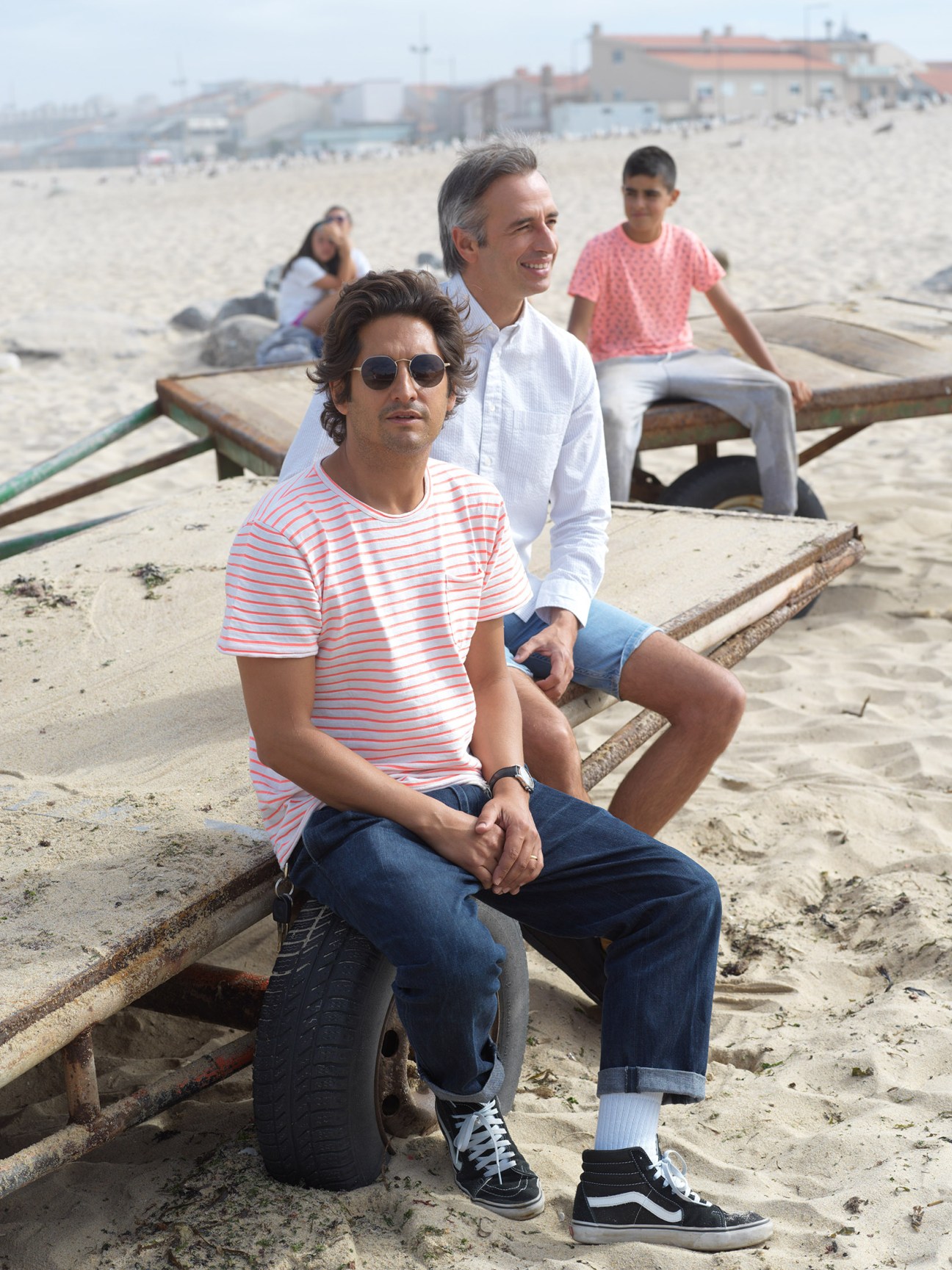 Two men sit on wooden boat timber on a sandy beach with coastal buildings visible behind them.
