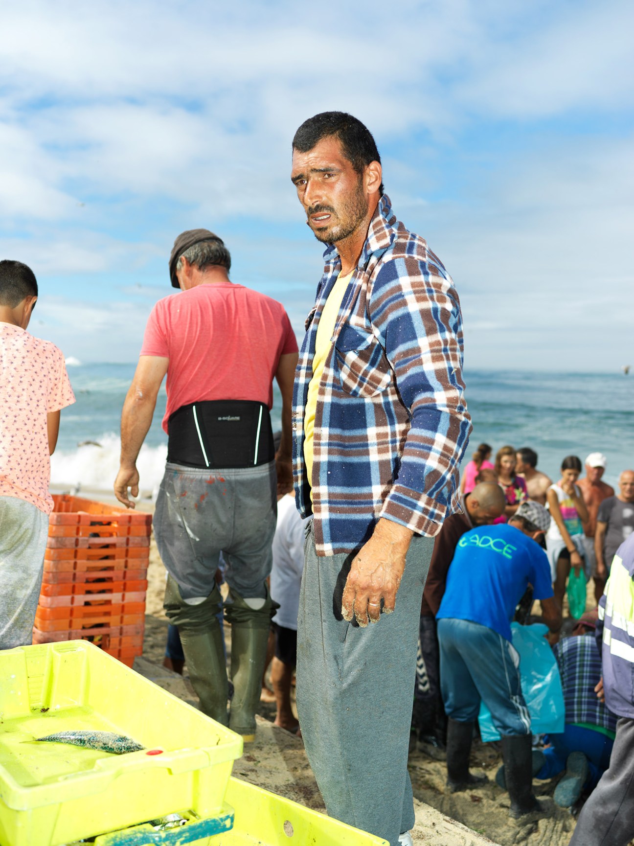Portuguese fisherman in plaid shirt stands on beach near yellow boat with catch.