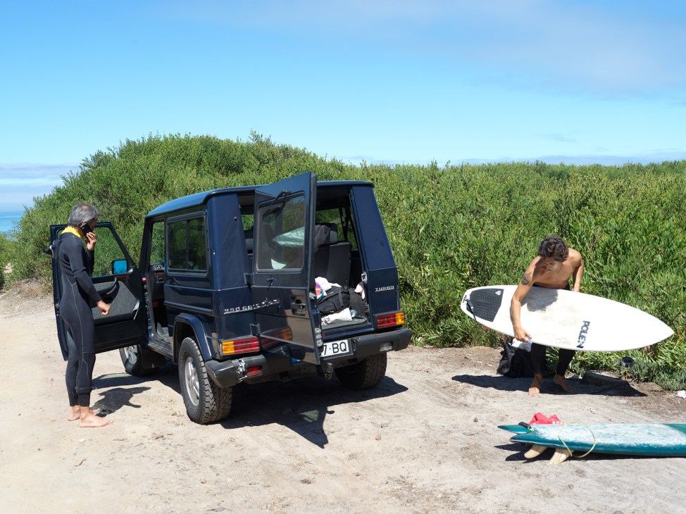 Two surfers with boards beside a black Mercedes G-Wagen on a sandy beach near shrubland.