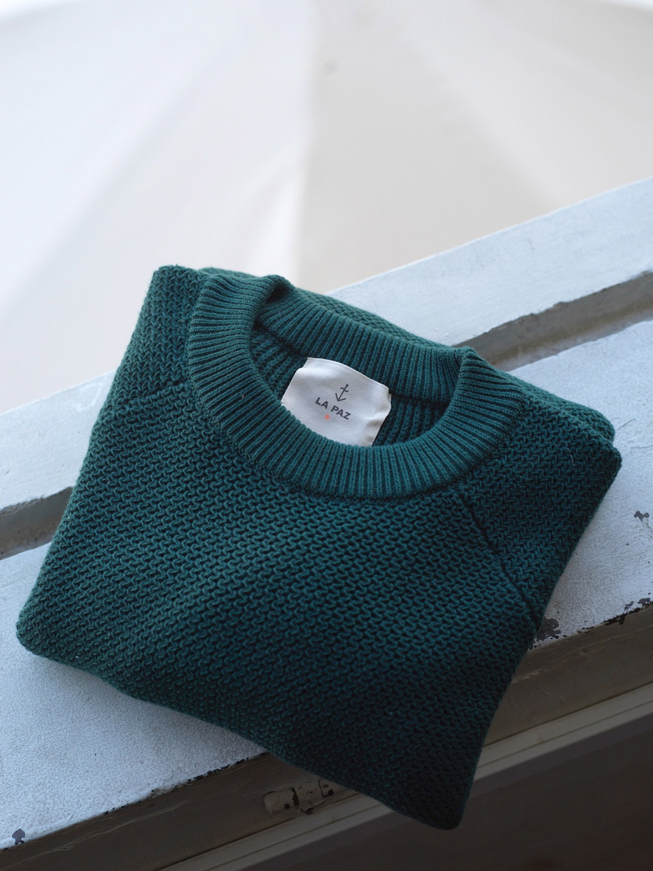 Folded teal cotton knit sweater with La Paz brand label on light blue surface.
